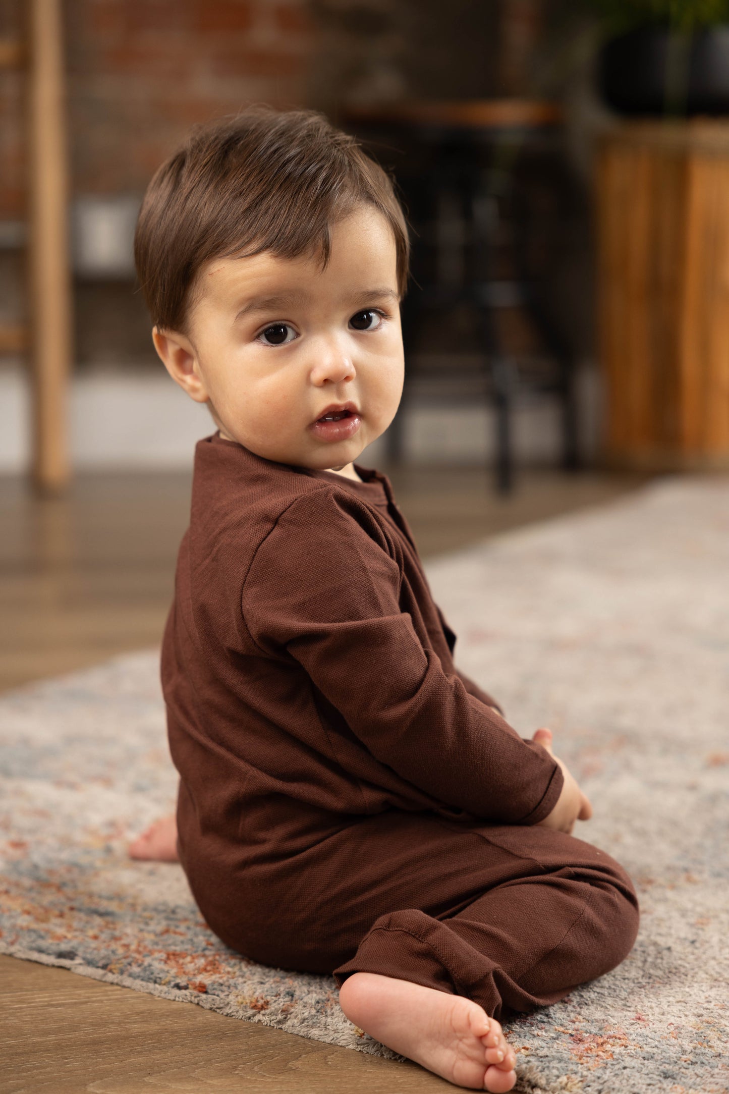 Baby in a brown outfit sitting on a rug with a blurred indoor background