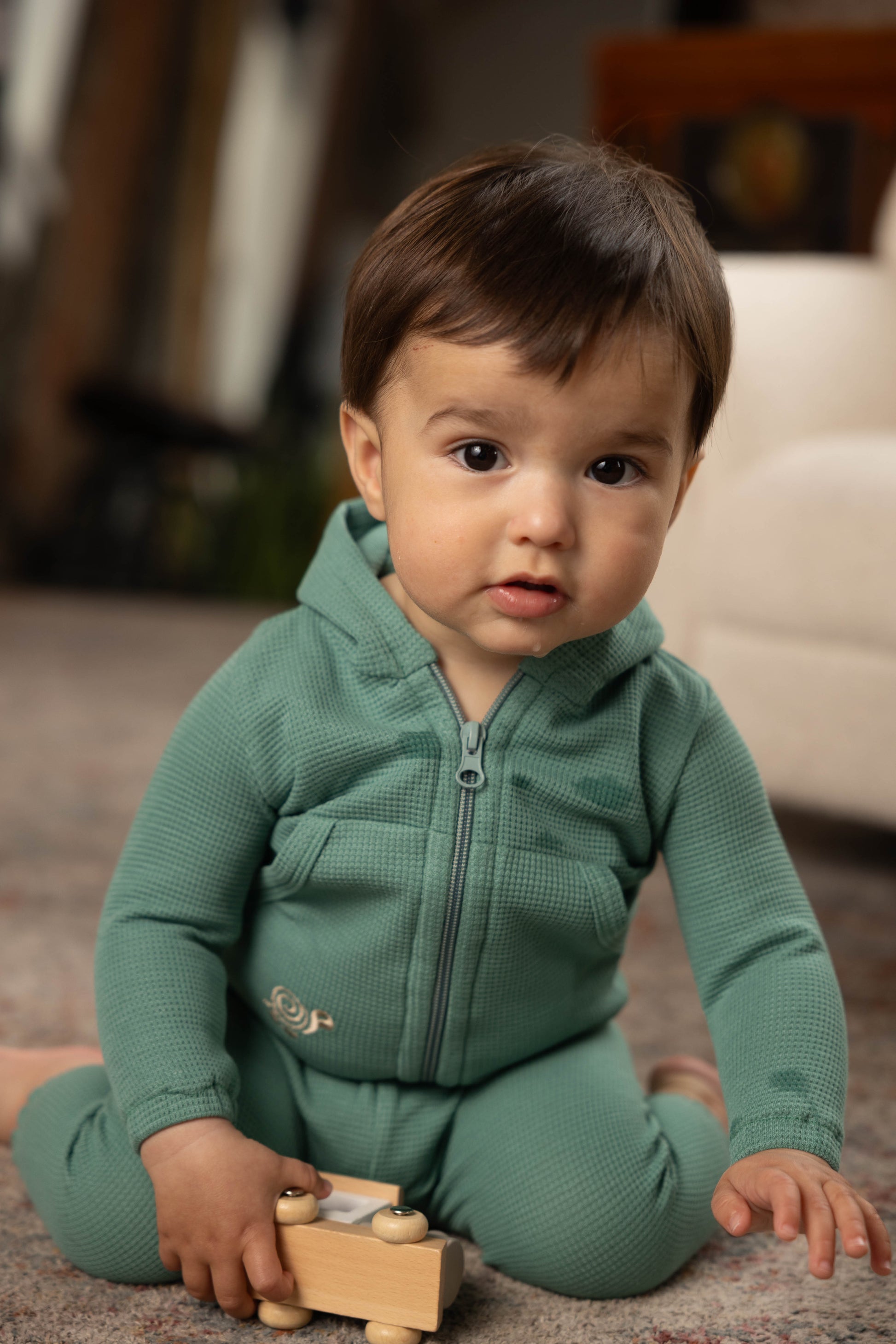 Baby in a green outfit sitting on the floor with a wooden toy.