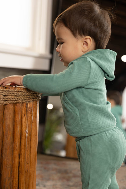 Child in a green outfit standing next to a wooden surface indoors.