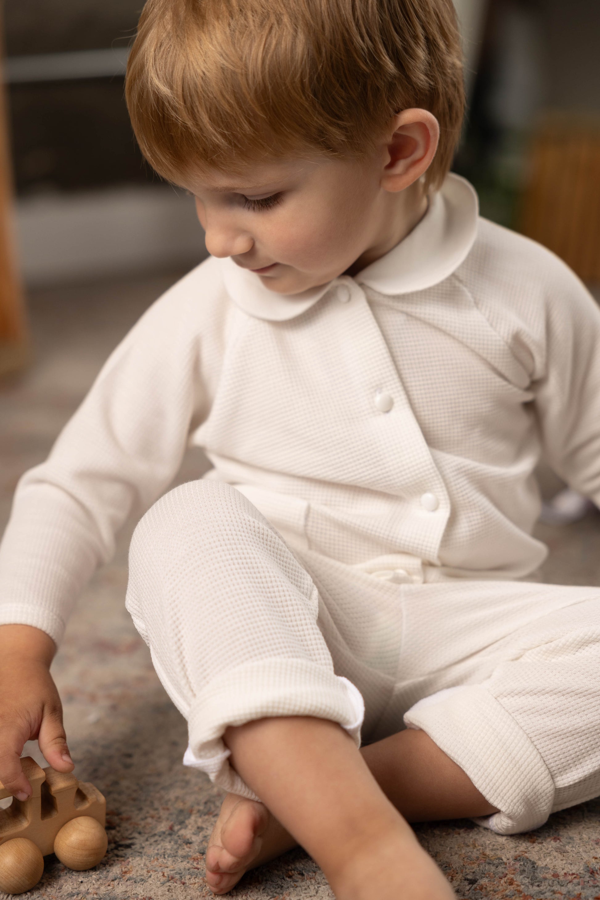 Child in a white waffle outfit playing with a wooden toy on a carpeted floor.