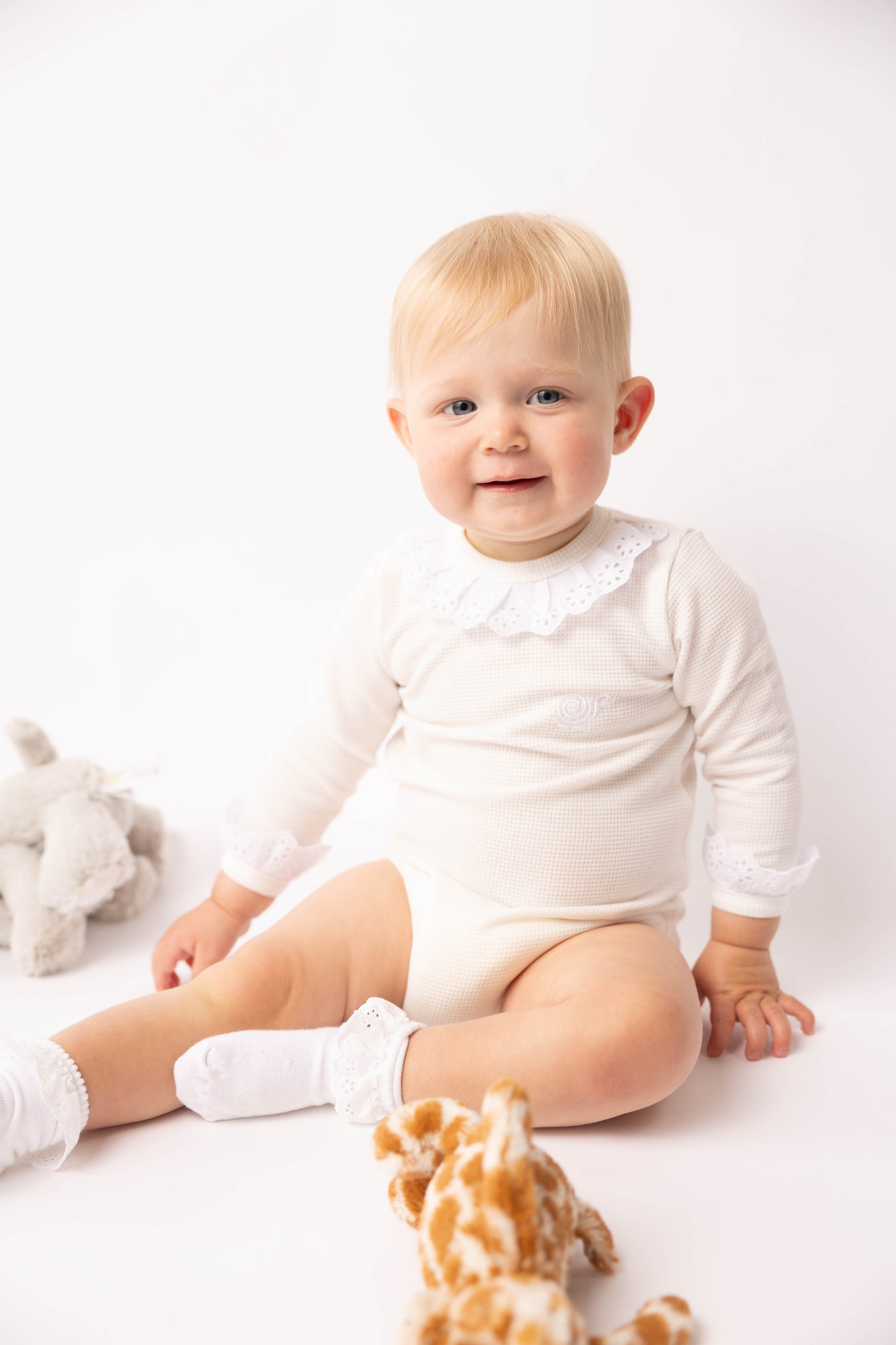 Baby in a white onesie sitting on a white surface with a toy giraffe and seashell.