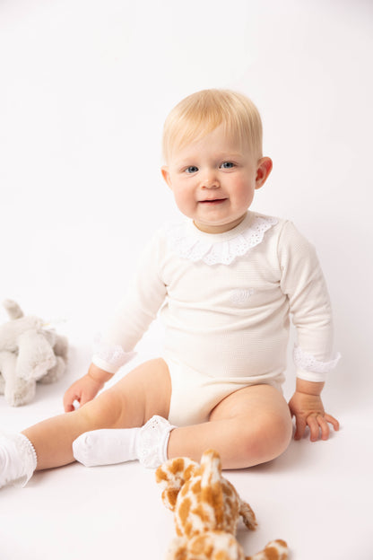 Baby in a white onesie sitting on a white surface with a toy giraffe and seashell.