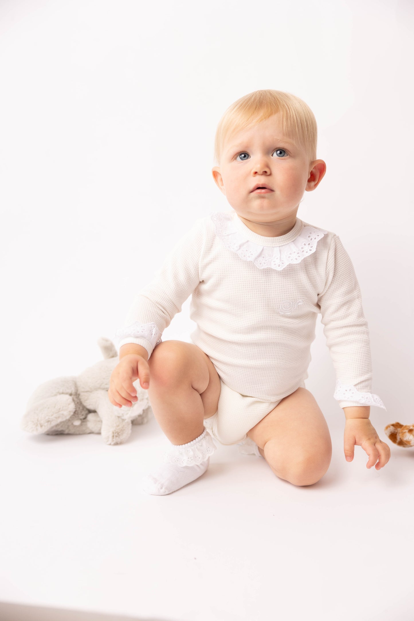 Baby in a white outfit sitting on a white surface with a stuffed toy.