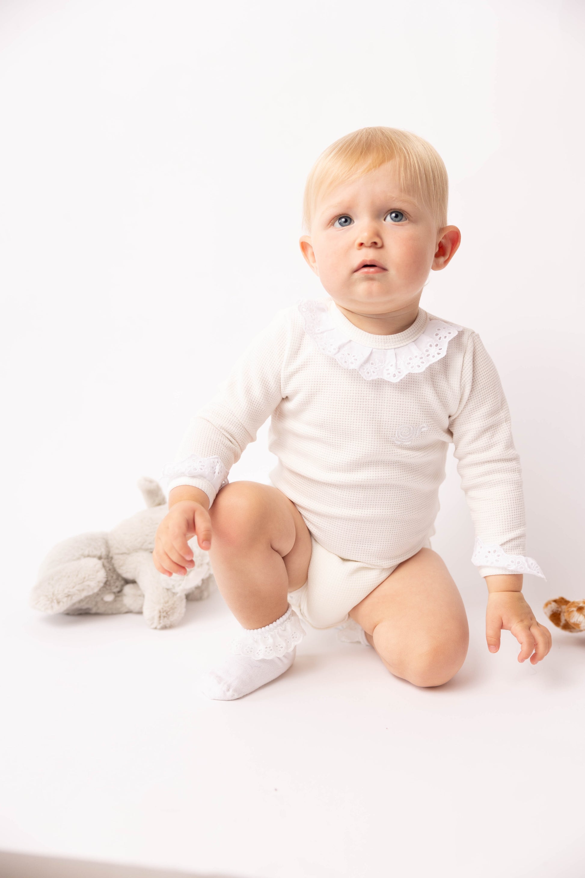Baby in a white outfit sitting on a white surface with a stuffed toy.